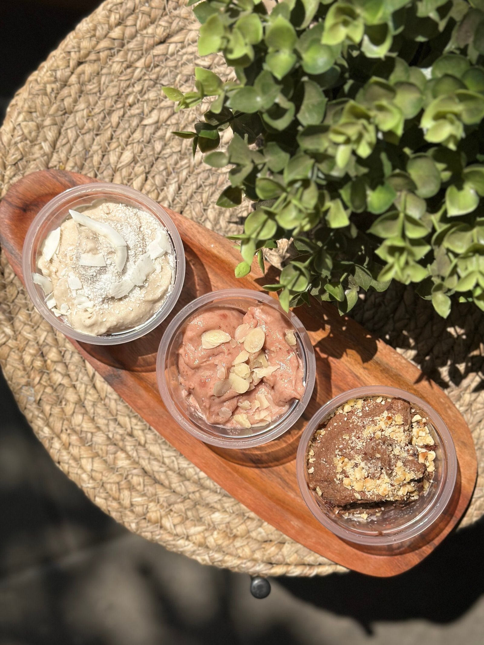 Top-down view of four cups of ice cream with toppings on a wooden board beside a green plant.