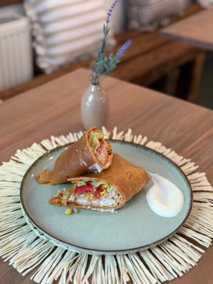 Sliced burrito filled with rice, peppers, and lettuce on a blue plate with a side of white sauce, set on a woven placemat. A small vase with lavender is blurred in the background.