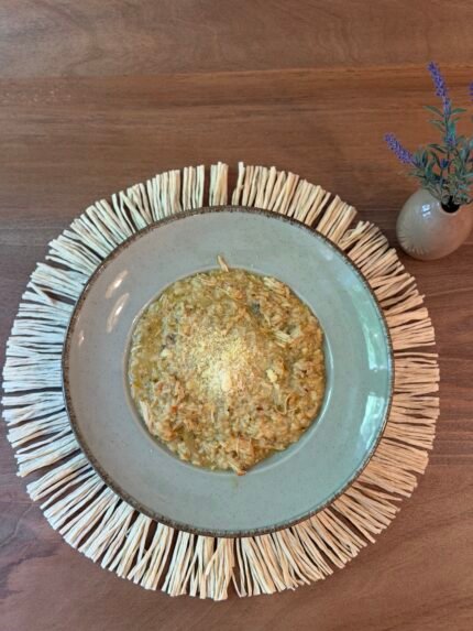Top-down view of a bowl of chunky vegetable soup on a ceramic plate, set on a woven straw placemat with a small vase of purple lavender on a wooden table.